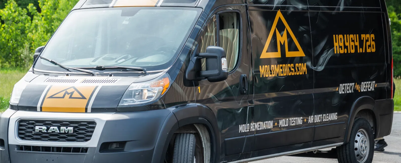 Black RAM van with Mold Medics branding parked on asphalt near green trees, featuring mold remediation services and contact info.