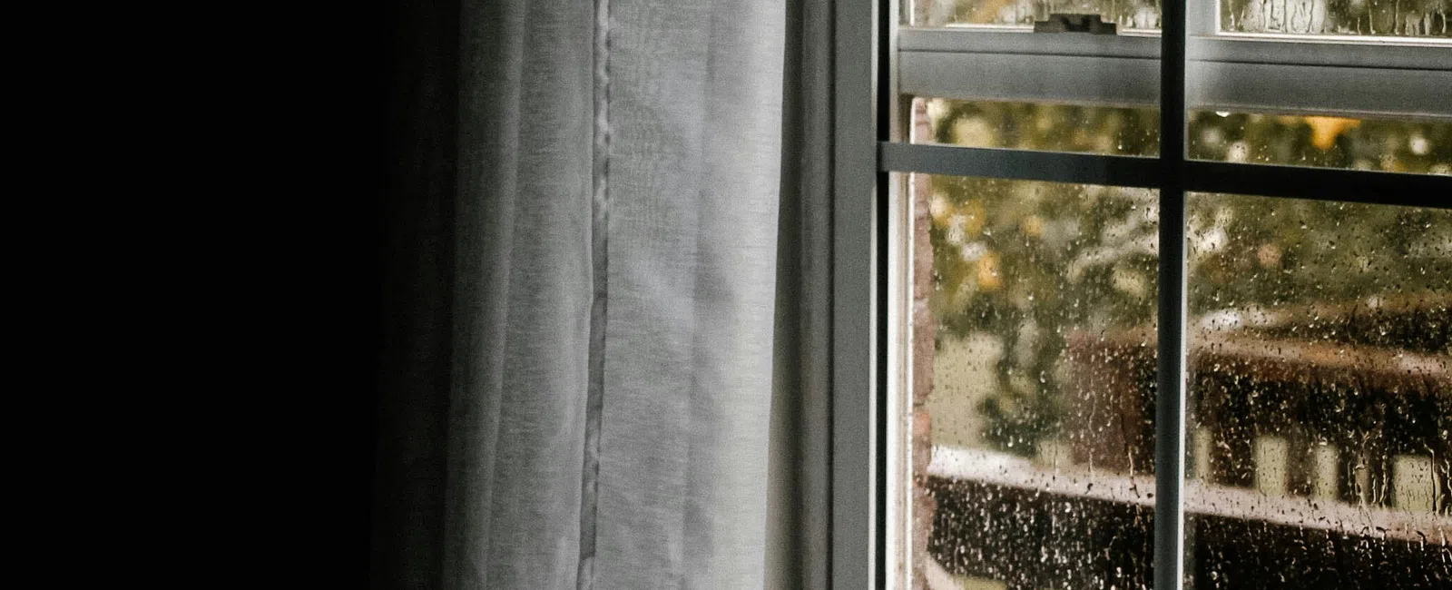 Rain droplets on a window with sheer white curtains partially drawn, showing an outdoor porch and greenery beyond.