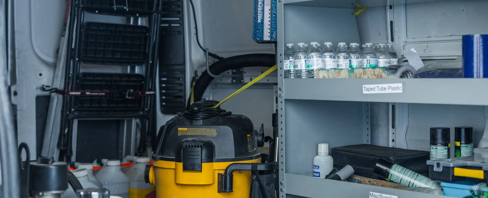 Organized van interior with cleaning equipment, tools, water bottles, and supplies on metal shelving and floor.