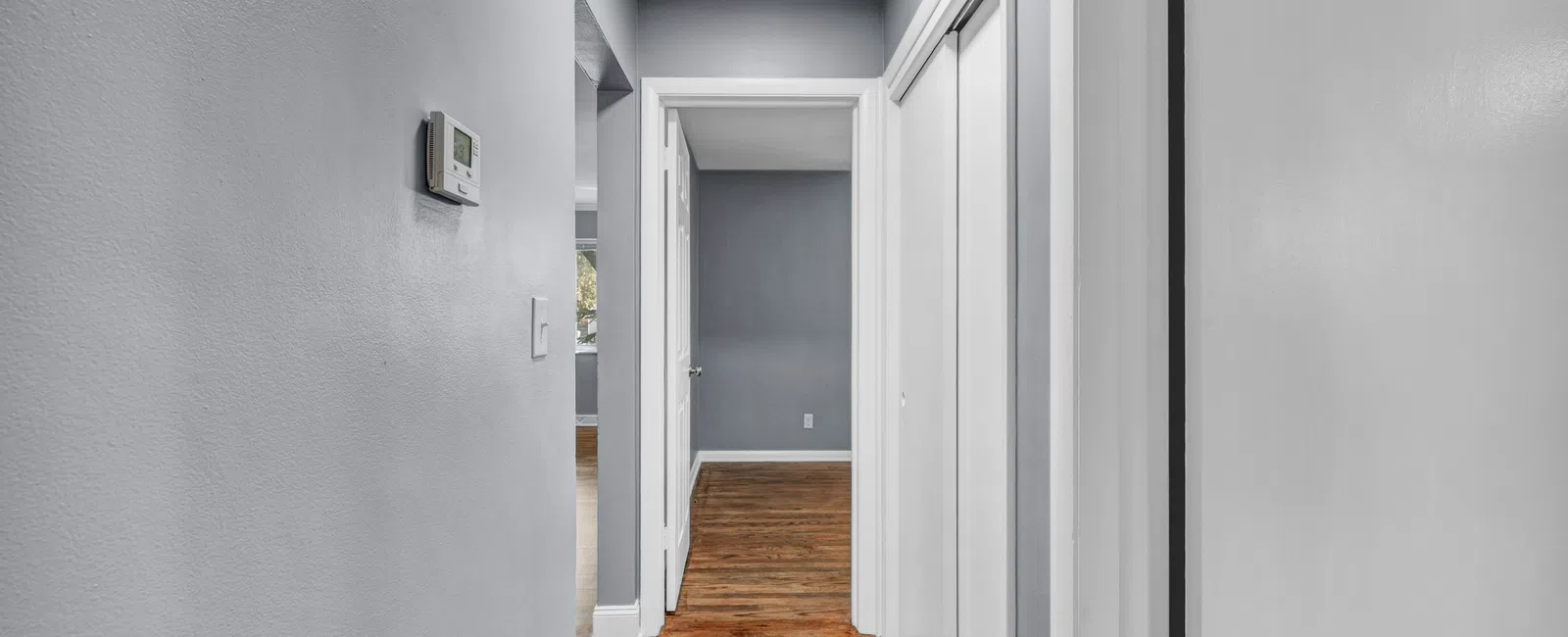Narrow hallway with gray walls, white doors, wooden floor, and ceiling light, leading to a gray-walled room.