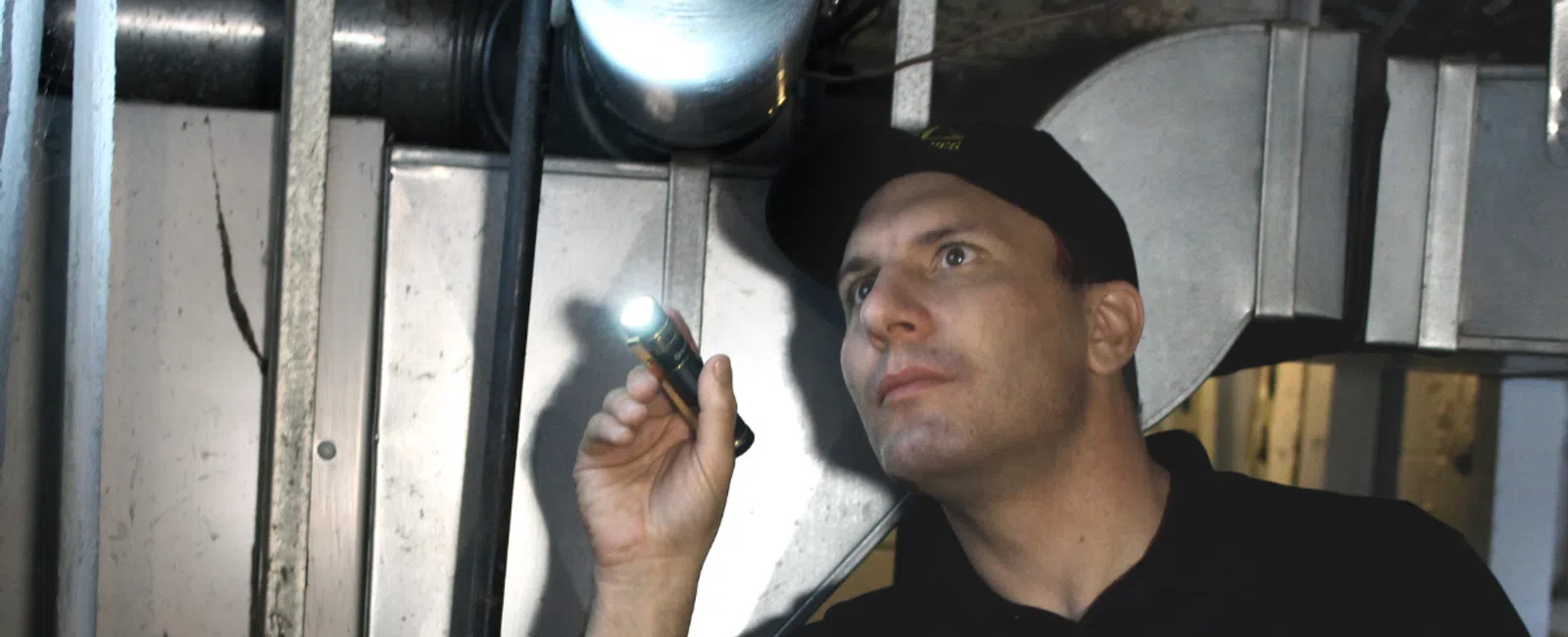 Technician in a Mold Medics polo inspects HVAC ducts with a flashlight in a dimly lit basement.