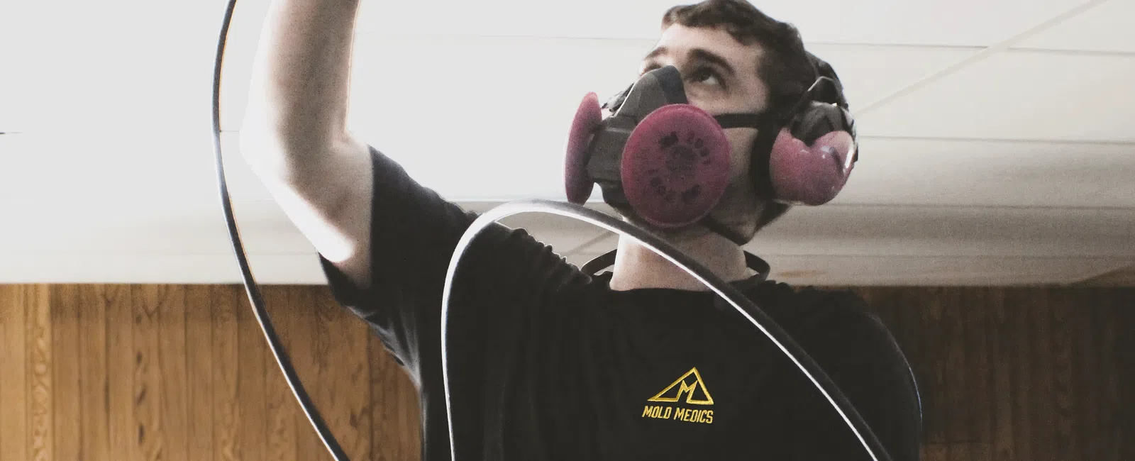 Technician wearing respirator and gloves inspecting ceiling panel with a hose in wood-paneled room.