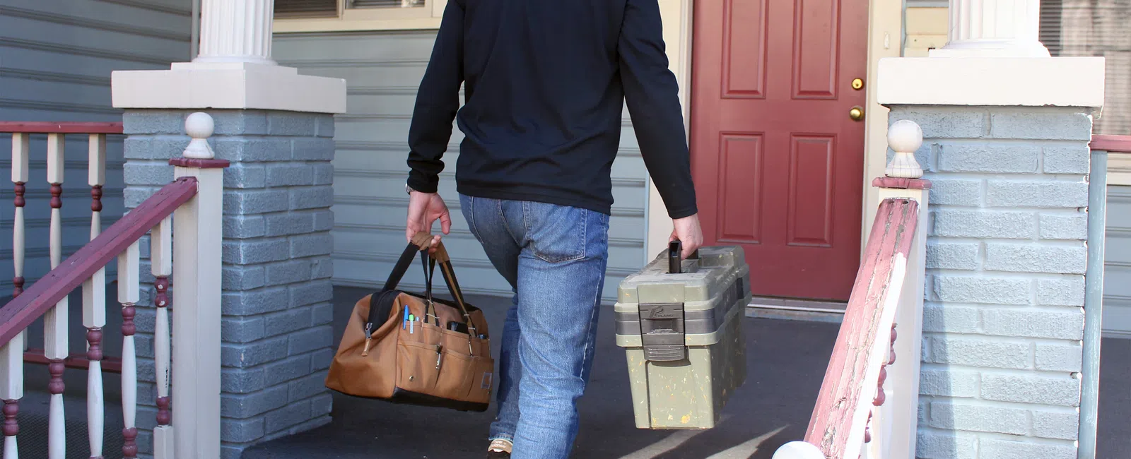 Technician carrying toolboxes walking up front porch stairs toward a red door at a residential house.