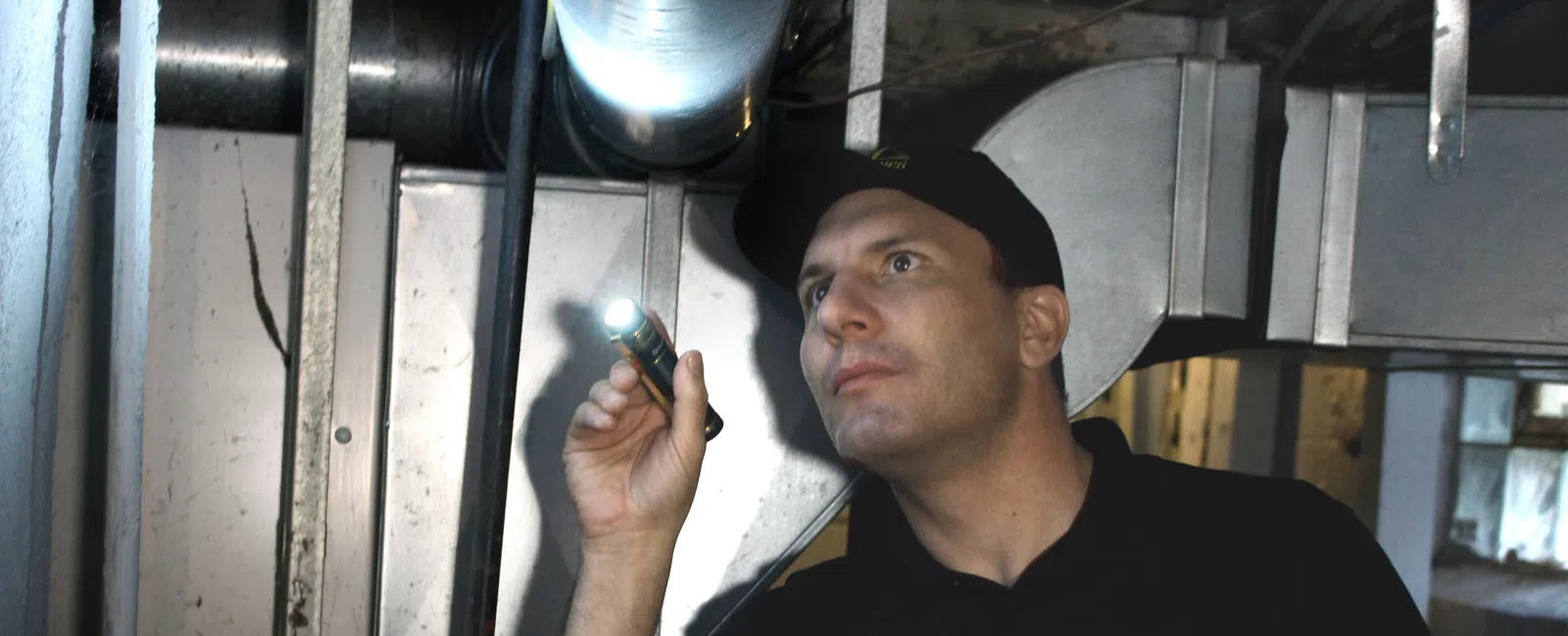 Technician in Mold Medics uniform inspecting ventilation ducts with flashlight in a dim basement.