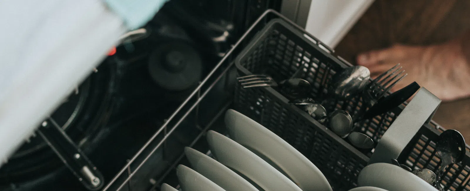Open dishwasher loaded with neatly arranged white plates, cutlery, and a glass cup in a modern kitchen.