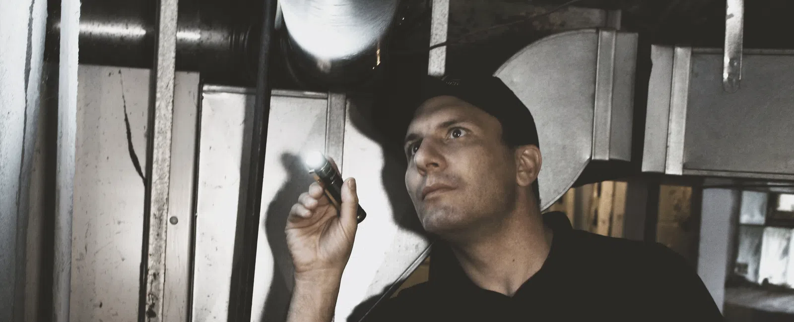 Technician inspecting HVAC system with flashlight in a basement wearing Mold Medics uniform and cap.