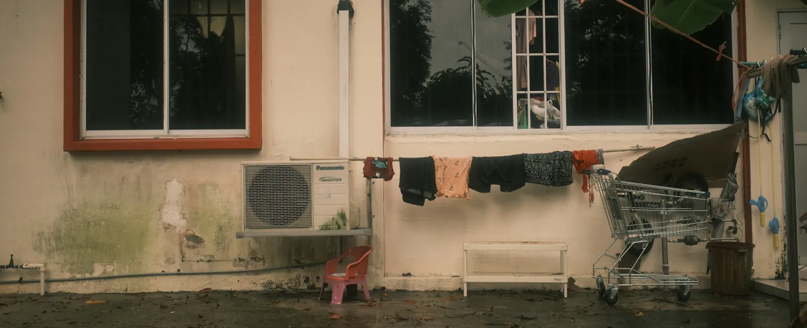 A weathered building with a clothesline, air conditioner, and a shopping cart on a rainy day.