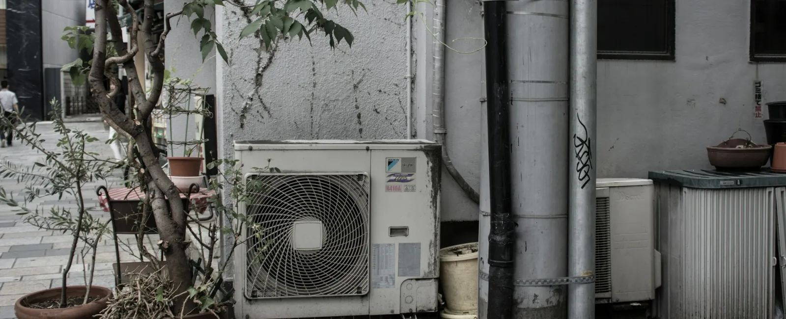 Air conditioning unit on a city street, surrounded by pots and urban elements, showcasing city life.