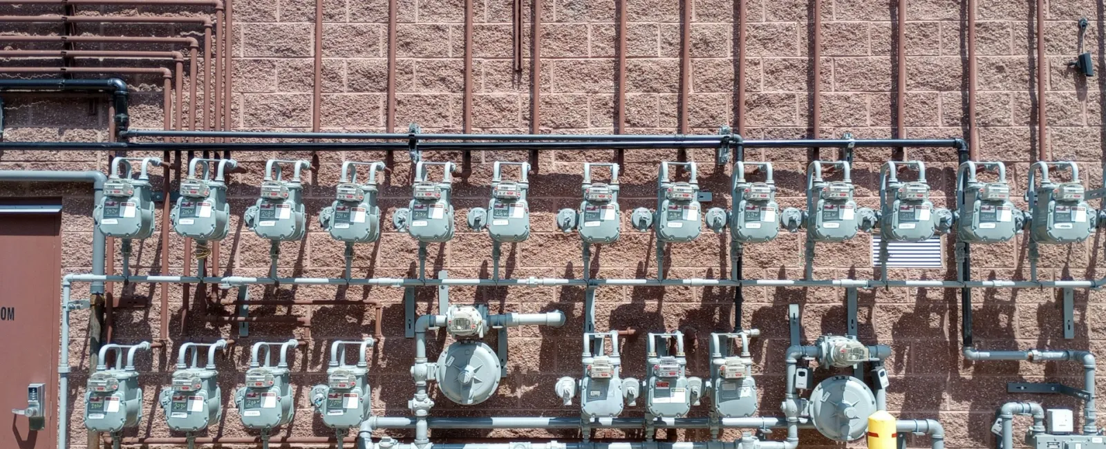 Wall of gas meters with pipes against a textured brick background in industrial setting.