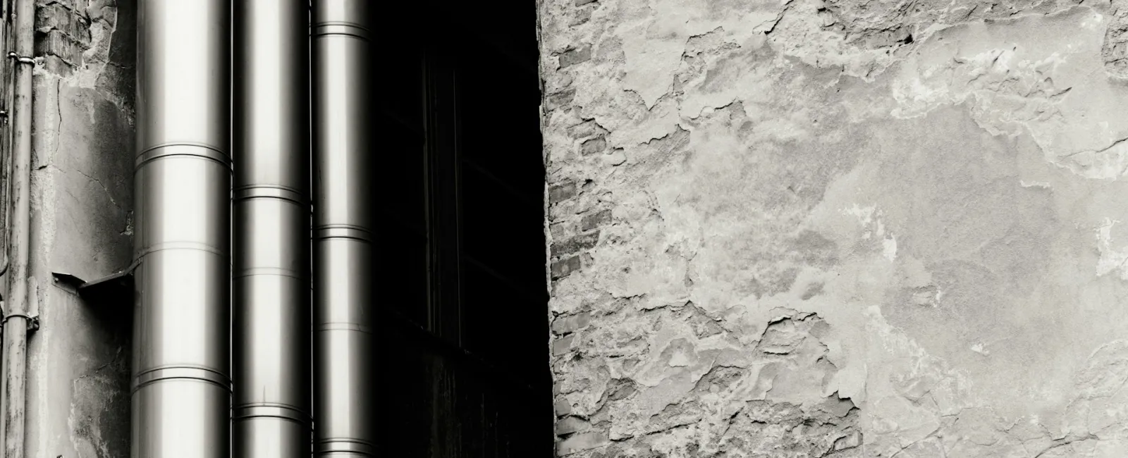 Black and white image capturing textured wall and metallic pipes juxtaposed.