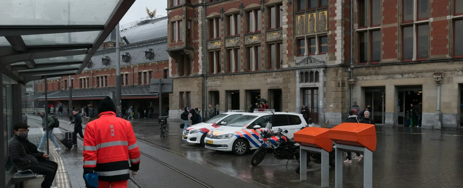 A wet street scene near a train station with police, pedestrians, and modern architecture.