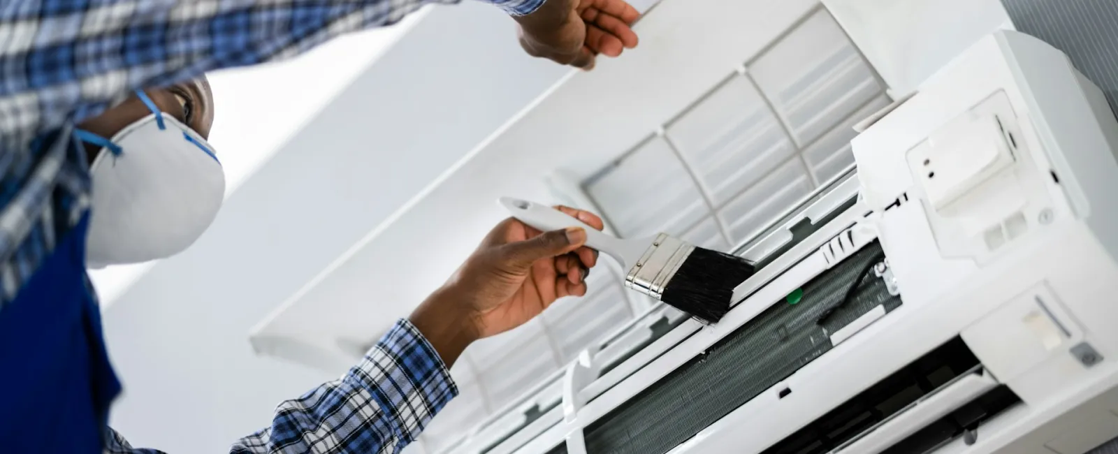 Technician cleaning air conditioning unit with a brush wearing a mask for safety.