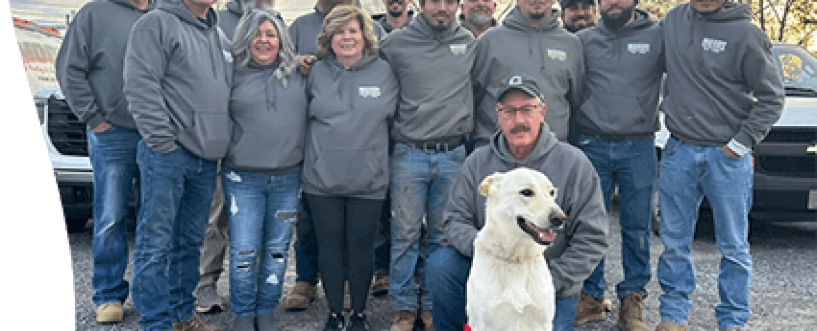 Team photo with a dog, showcasing camaraderie in gray uniforms amidst trucks and a sunset sky.