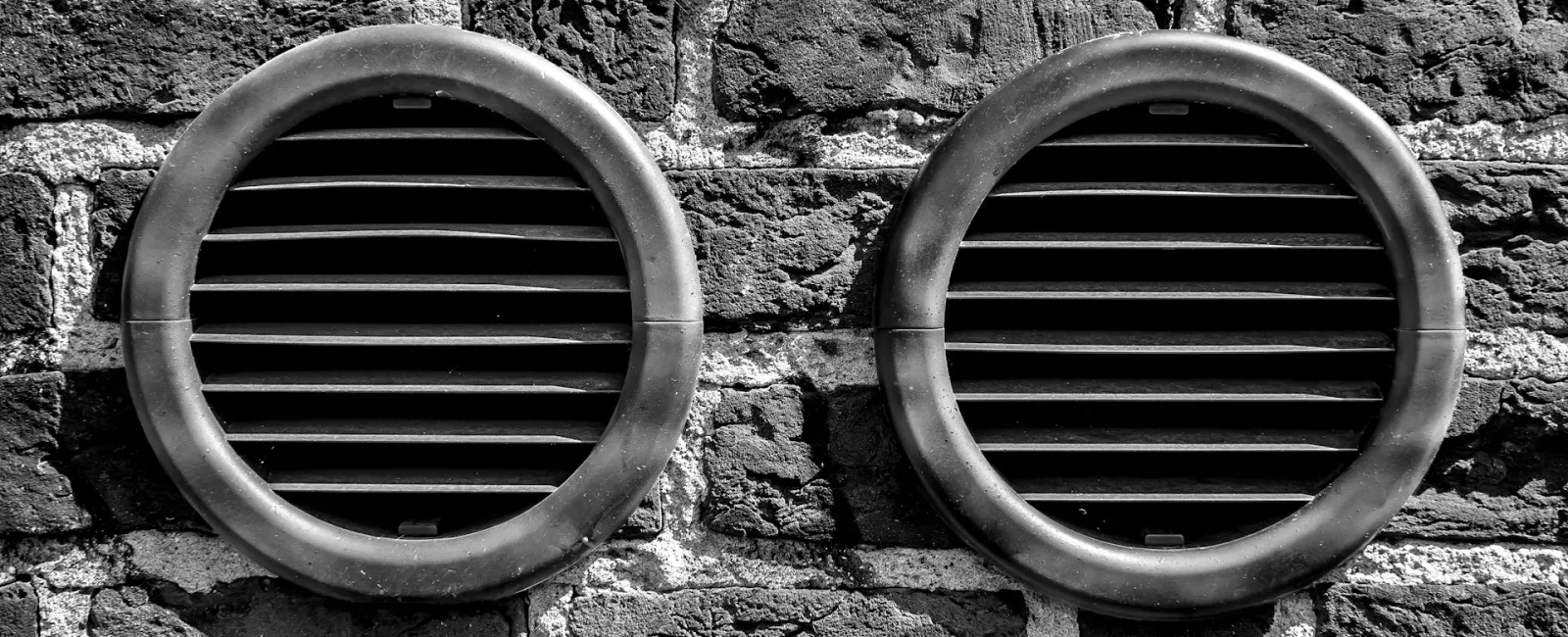 Black and white photo of two circular wall vents with metal grilles on a textured brick wall.