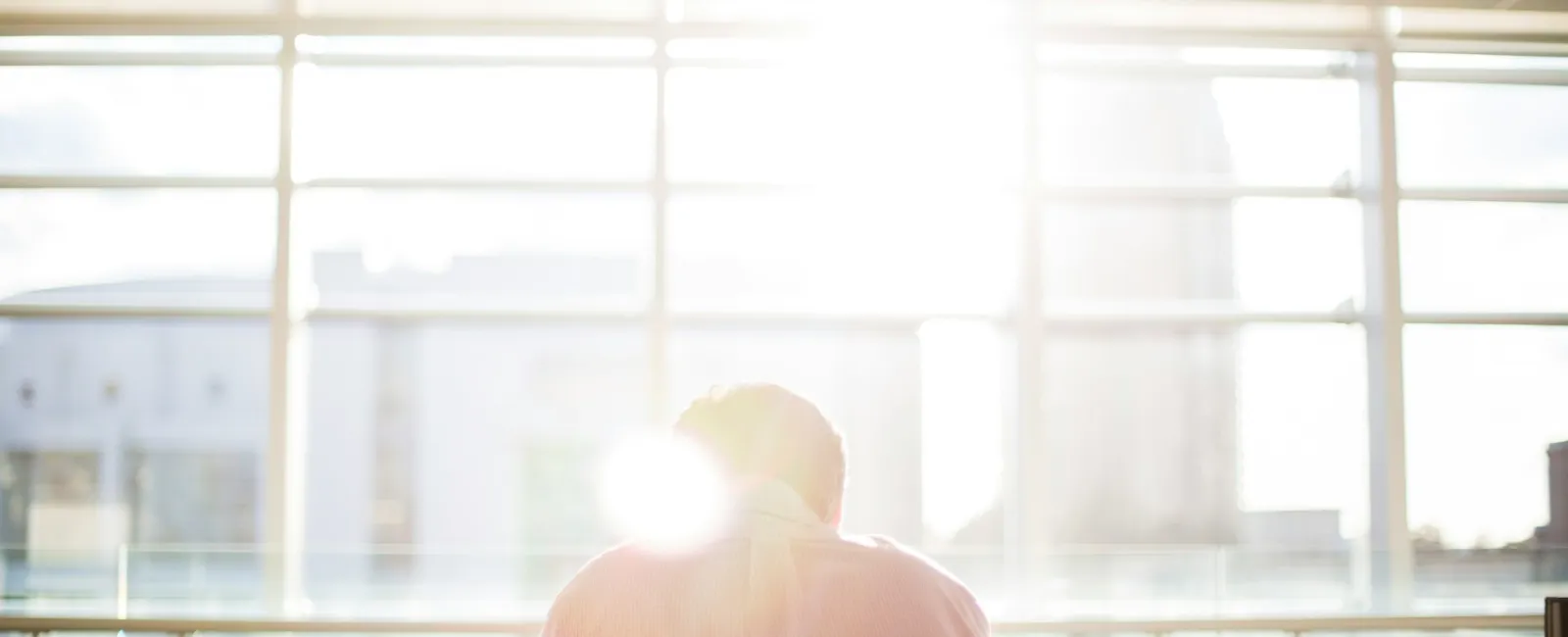 Silhouette of a person sitting at a desk, sunlight streaming through large windows in a modern office.