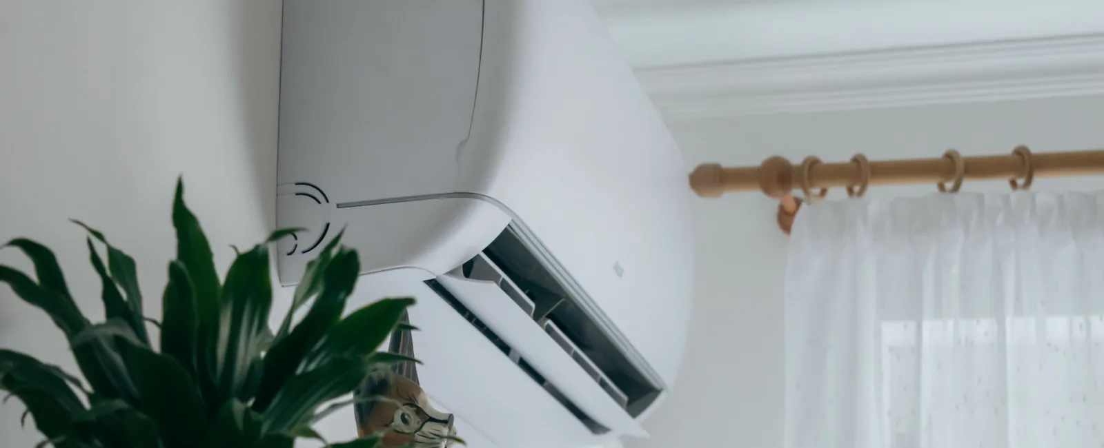 Modern air conditioner mounted on the wall beside a green potted plant in a bright room.