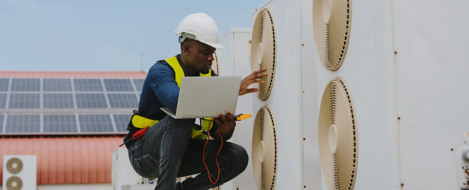 Technician in safety gear inspecting large HVAC units with a laptop and electrical testing tools outdoors on a rooftop.