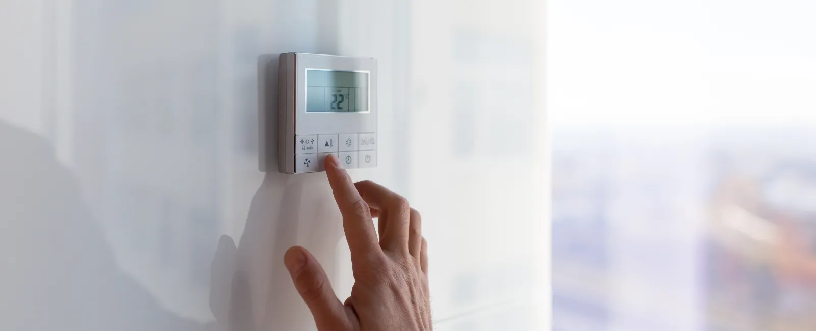 Hand adjusting a digital thermostat mounted on a white wall with soft natural light.