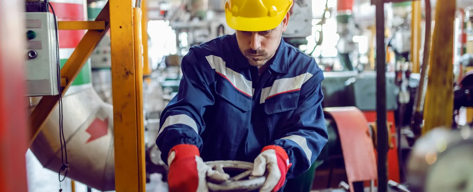 Industrial worker in protective gear adjusting valve in busy factory with machinery and pipes.