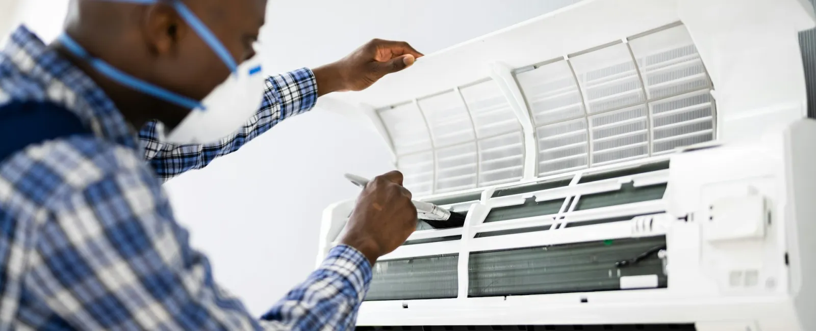 Technician wearing mask cleaning and servicing a white wall-mounted air conditioning unit indoors.