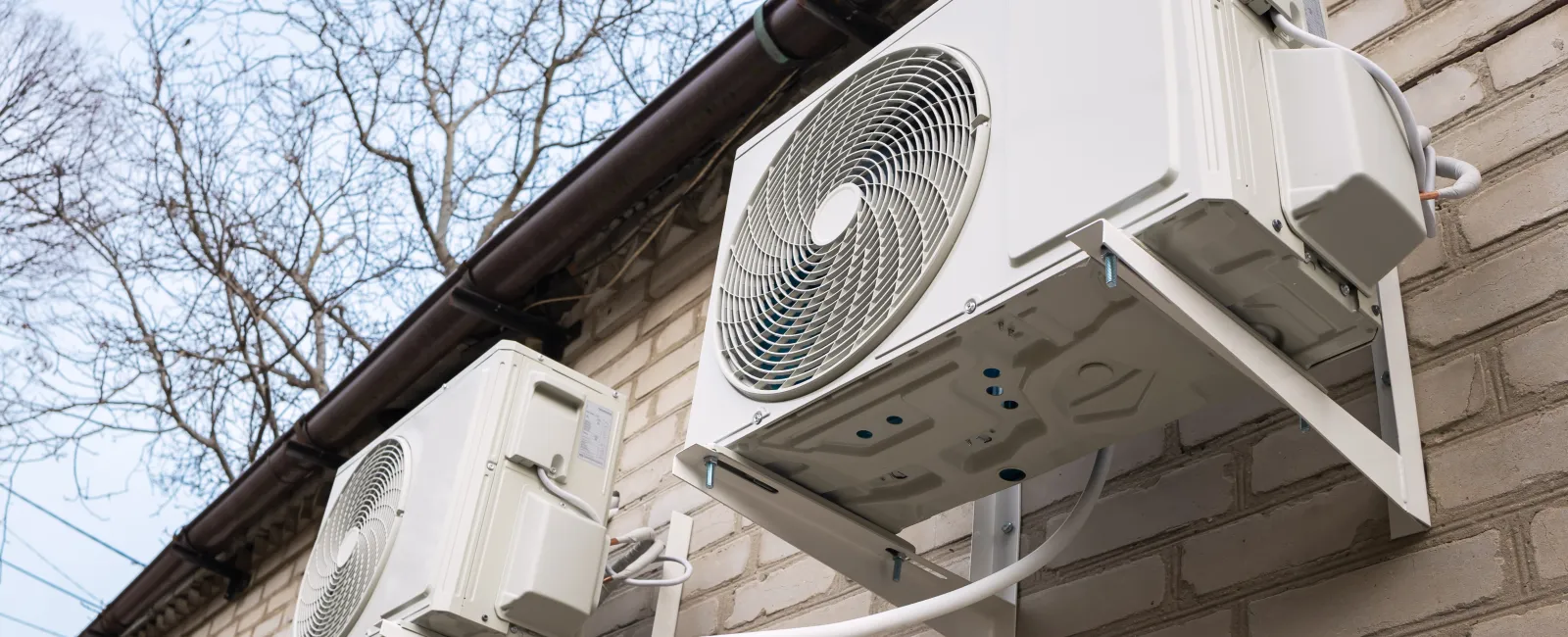 Two white outdoor air conditioning units mounted on a brick wall under a gutter with bare trees in the background.
