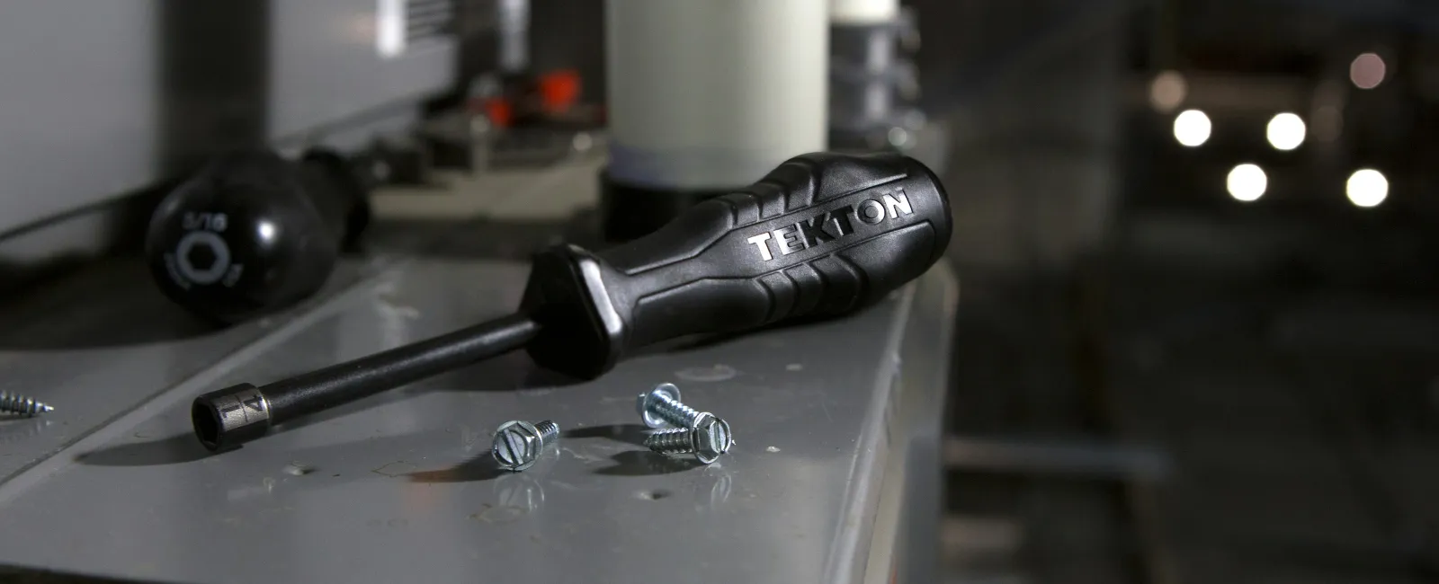 Tekton screwdriver and screws on a metal surface with blurred background and lighting bokeh.
