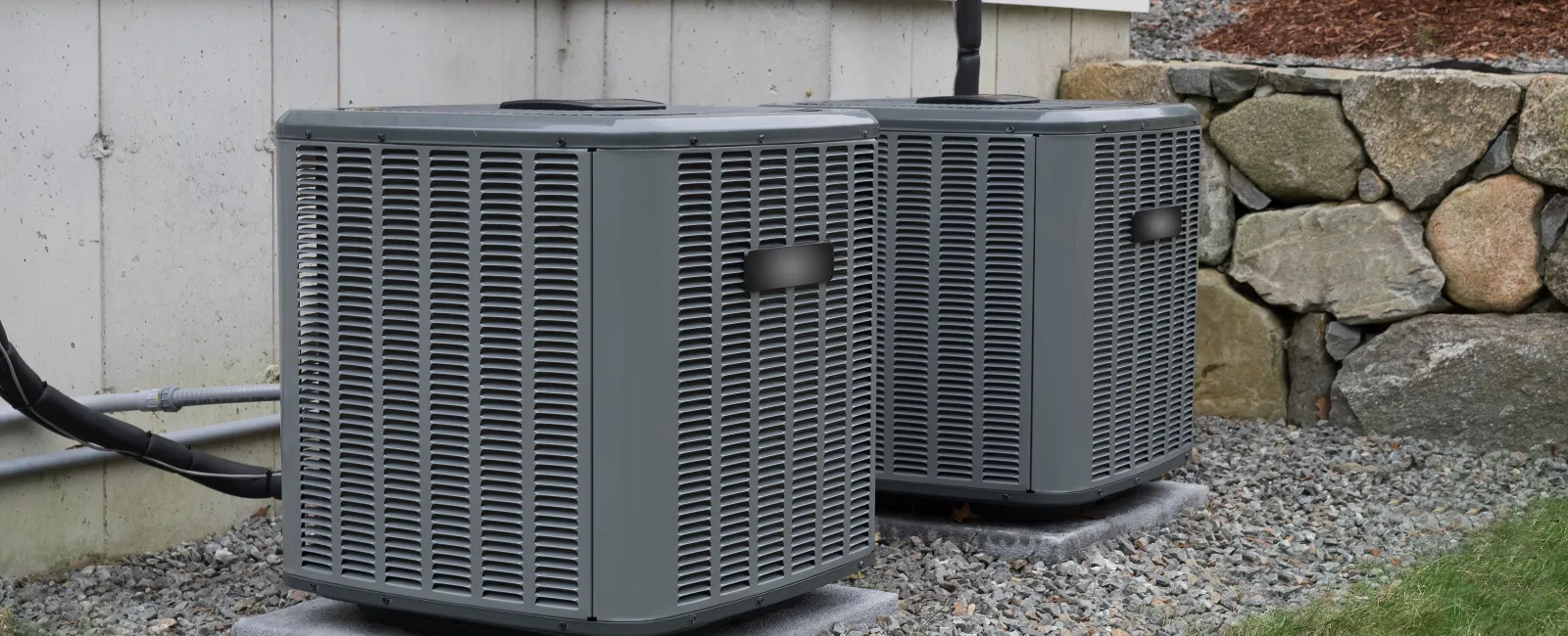 Two modern gray air conditioning units placed beside a house on a gravel base.