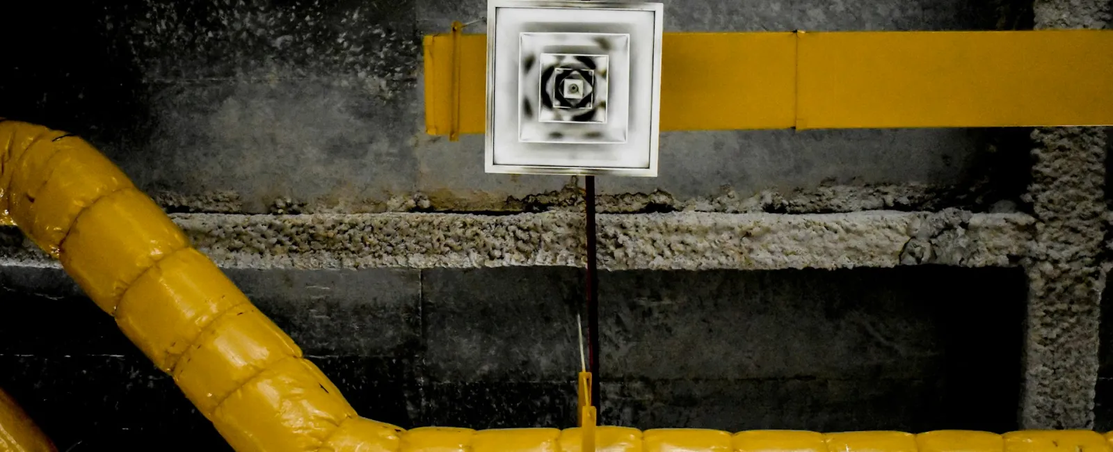 Yellow insulated pipes against a dark industrial wall with a white square vent in the center