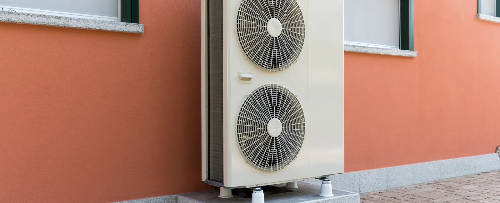 Outdoor heat pump unit installed beside an orange wall with windows.