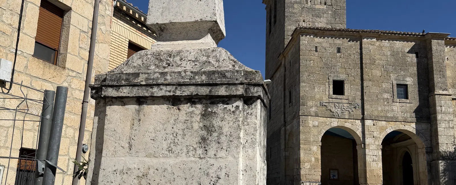 Historic stone fountain with rooster statue and coat of arms near old church under blue sky.