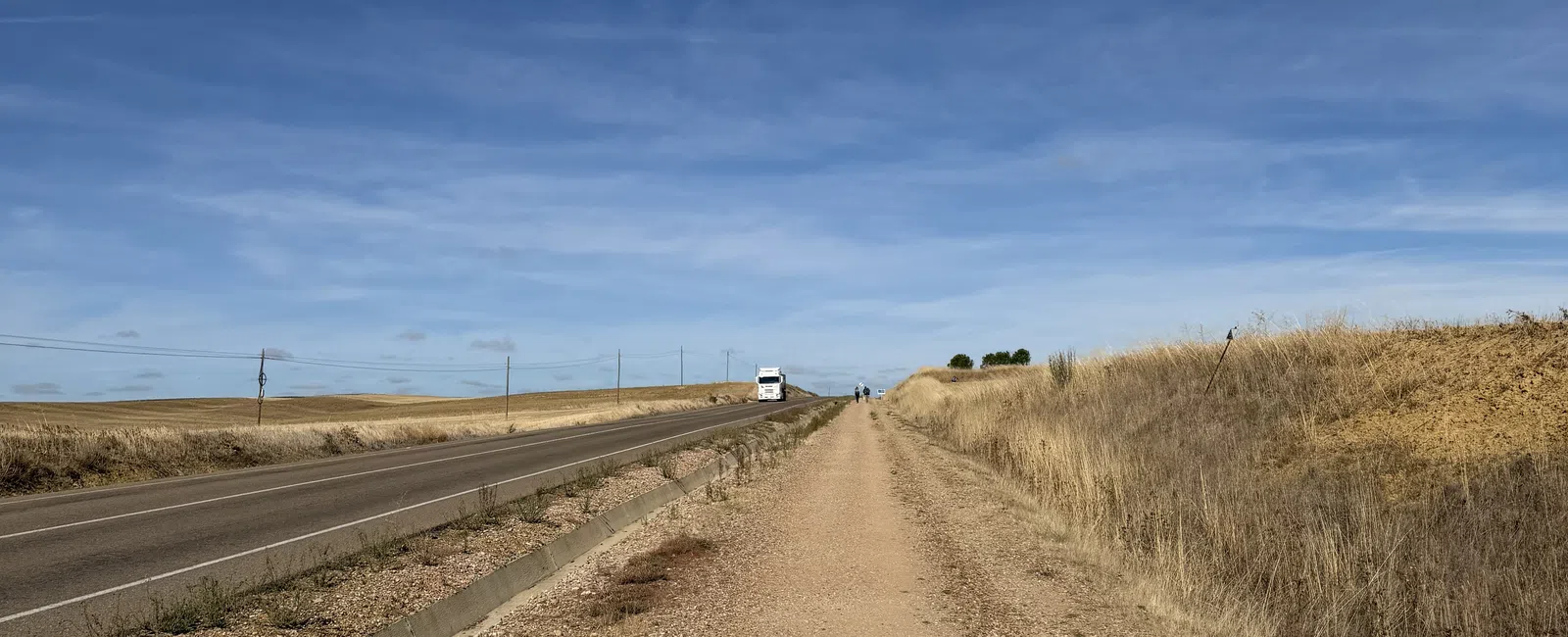 Gravel walking path parallel to a rural road under a clear blue sky with dry grass on the sides.