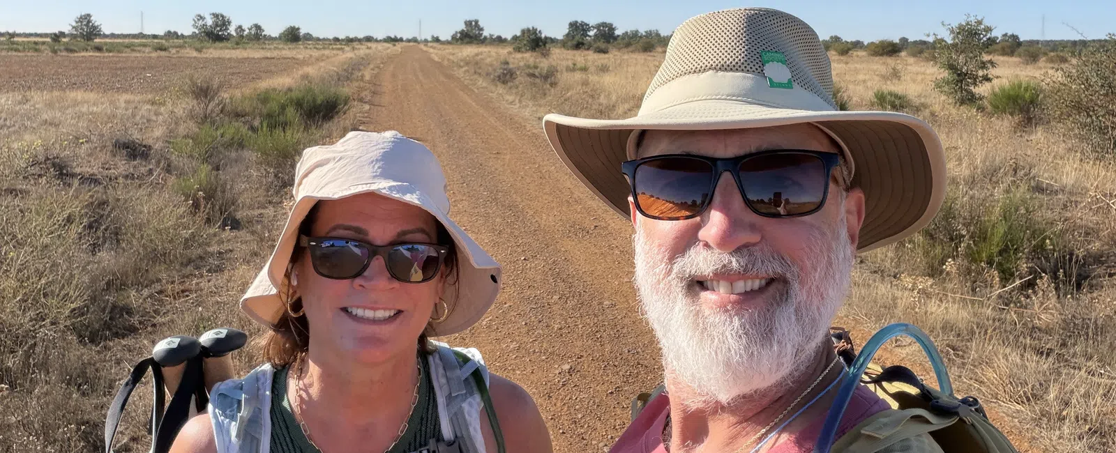 Smiling hikers wearing hats and backpacks on a dry dirt trail under a clear blue sky in a rural landscape