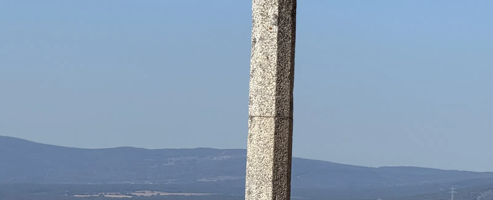 Stone cross monument with small stones on its base overlooking a cityscape under clear blue sky.