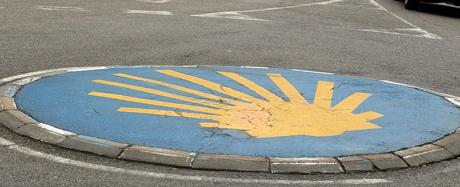 Roundabout with a yellow scallop shell symbol on a blue background in a suburban street with a white house and car nearby.