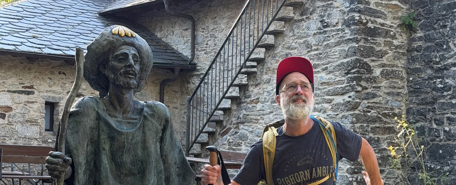 Man in hiking gear posing with bronze statue of pilgrim holding walking stick outside stone building with stairs.