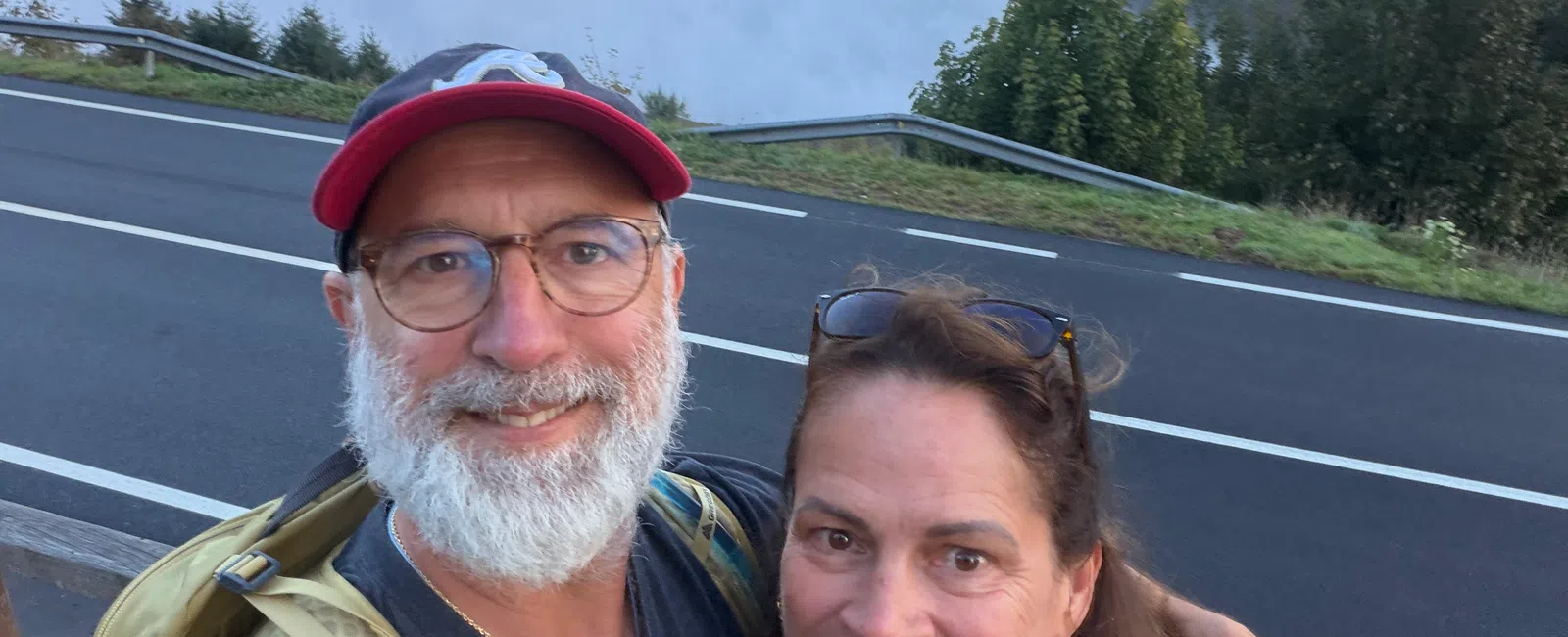 Smiling couple selfie with backpacks on mountain road overlooking fog-filled valley and cloudy sky.