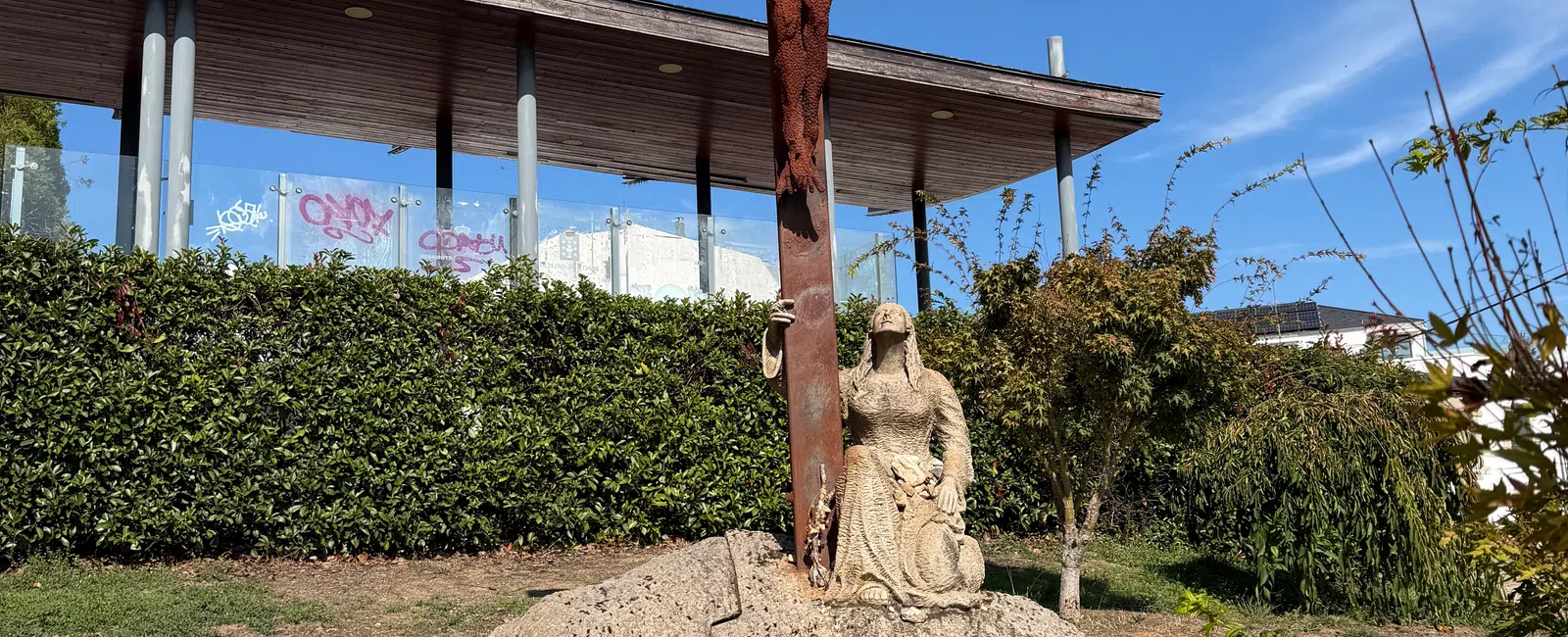 Rusty metal crucifix with Jesus and stone sculpture of kneeling woman on a rock under a clear blue sky