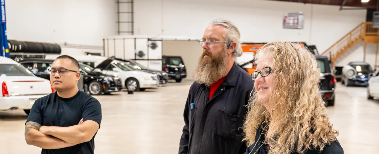 Three people standing and talking in a spacious car workshop with vehicles in the background.