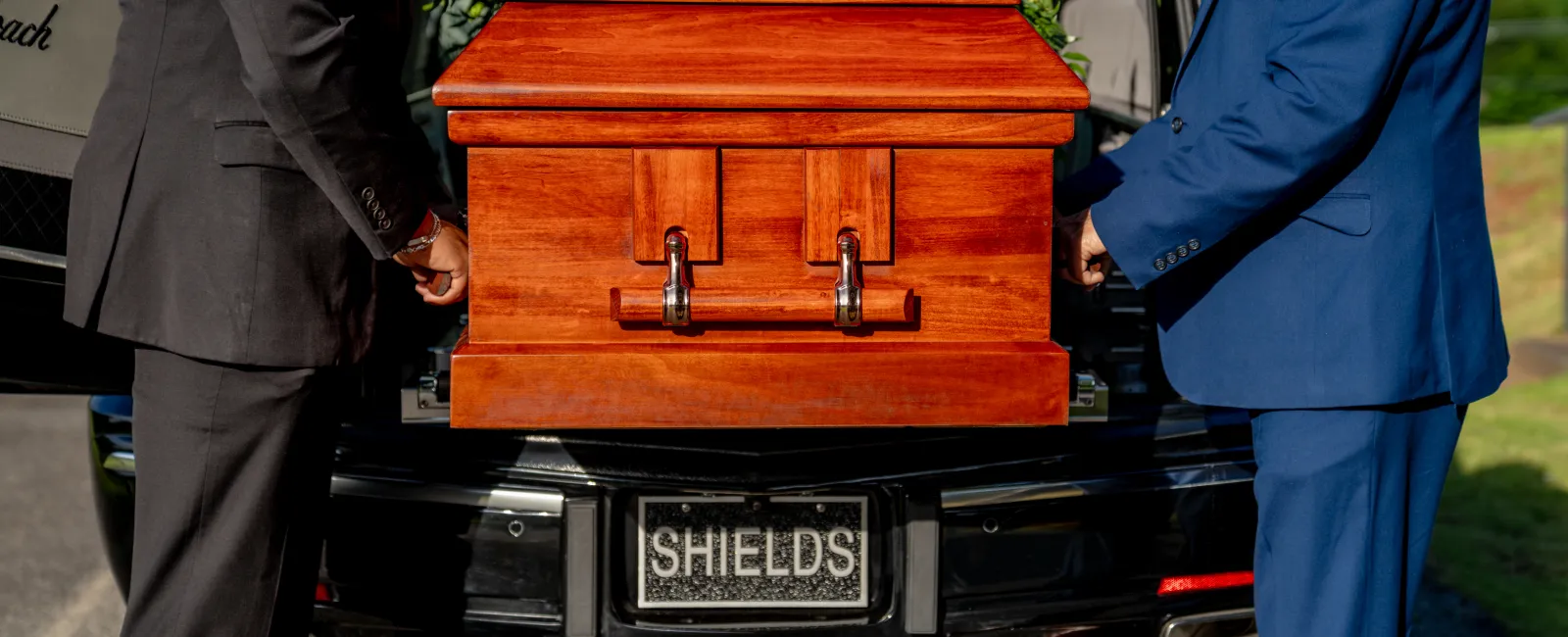 Two men in suits loading a polished wooden coffin into a black hearse with a floral arrangement on top