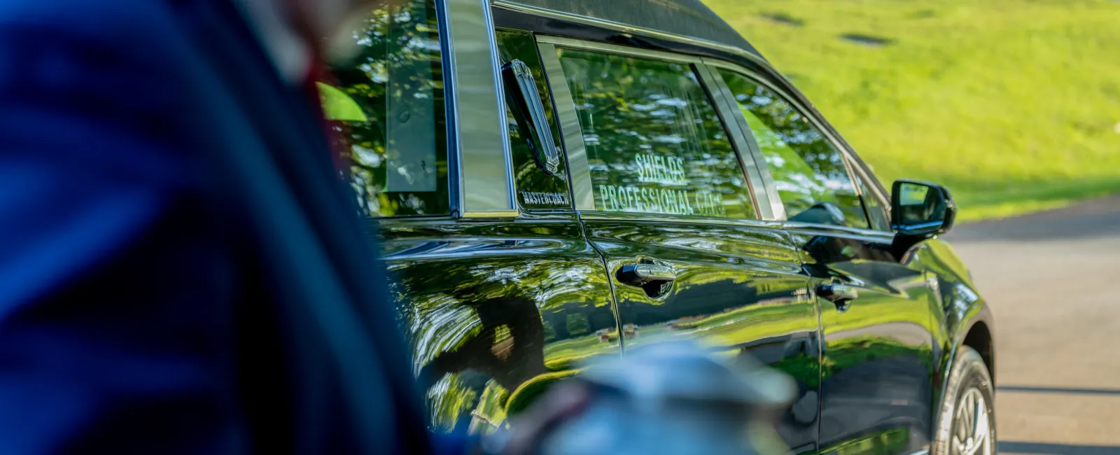 Black professional funeral car parked on a sunny day with an out-of-focus person holding a silver urn nearby