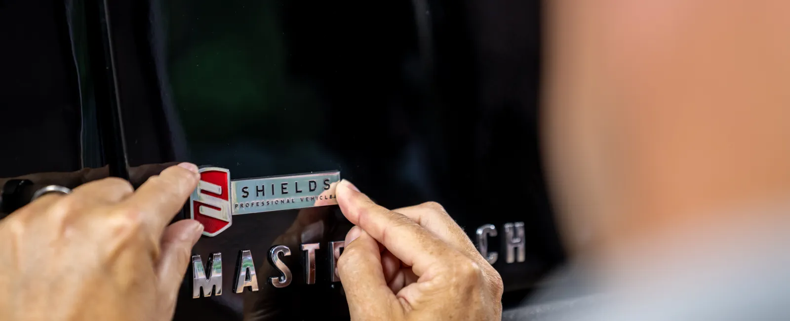 Close-up of hands applying a Shields Professional Vehicles emblem badge on a black Master Coach vehicle exterior.