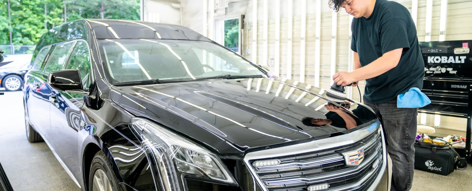 Man detailing a shiny black Cadillac sedan inside a bright garage with tools and equipment in the background
