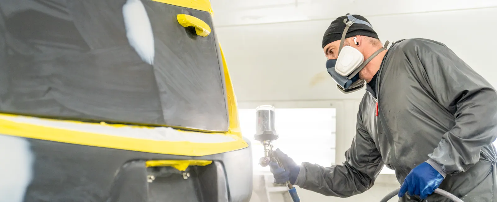 Worker in protective gear spray painting a car in a professional auto body shop with bright lighting.