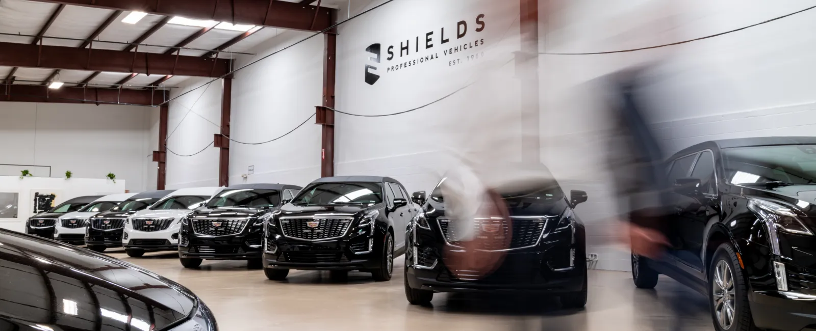 Line of luxury black and white Cadillac vehicles inside a spacious Shields Professional Vehicles showroom with blurred figures walking
