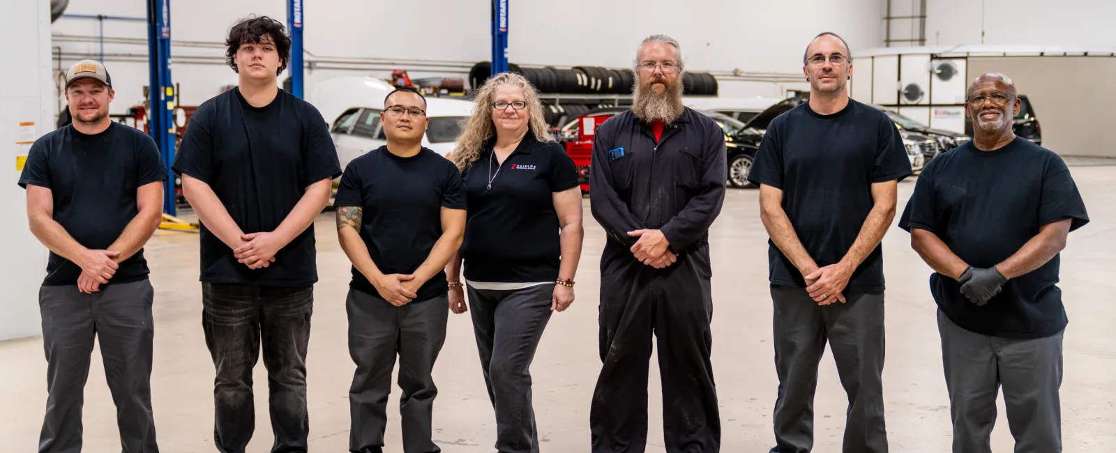 Seven auto mechanics standing in a spacious garage with car lifts and vehicles in the background.