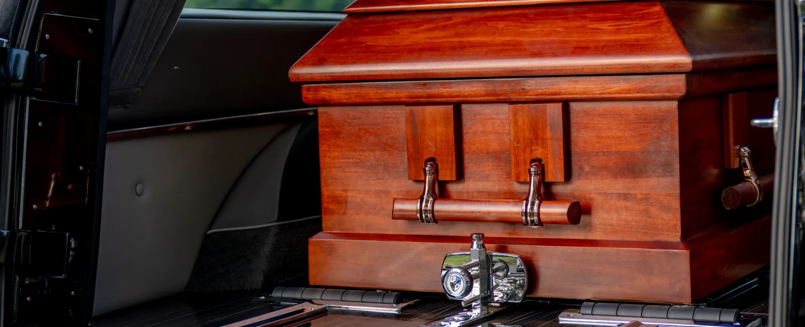 Closed wooden casket with flowers inside a black hearse with open rear door under daylight