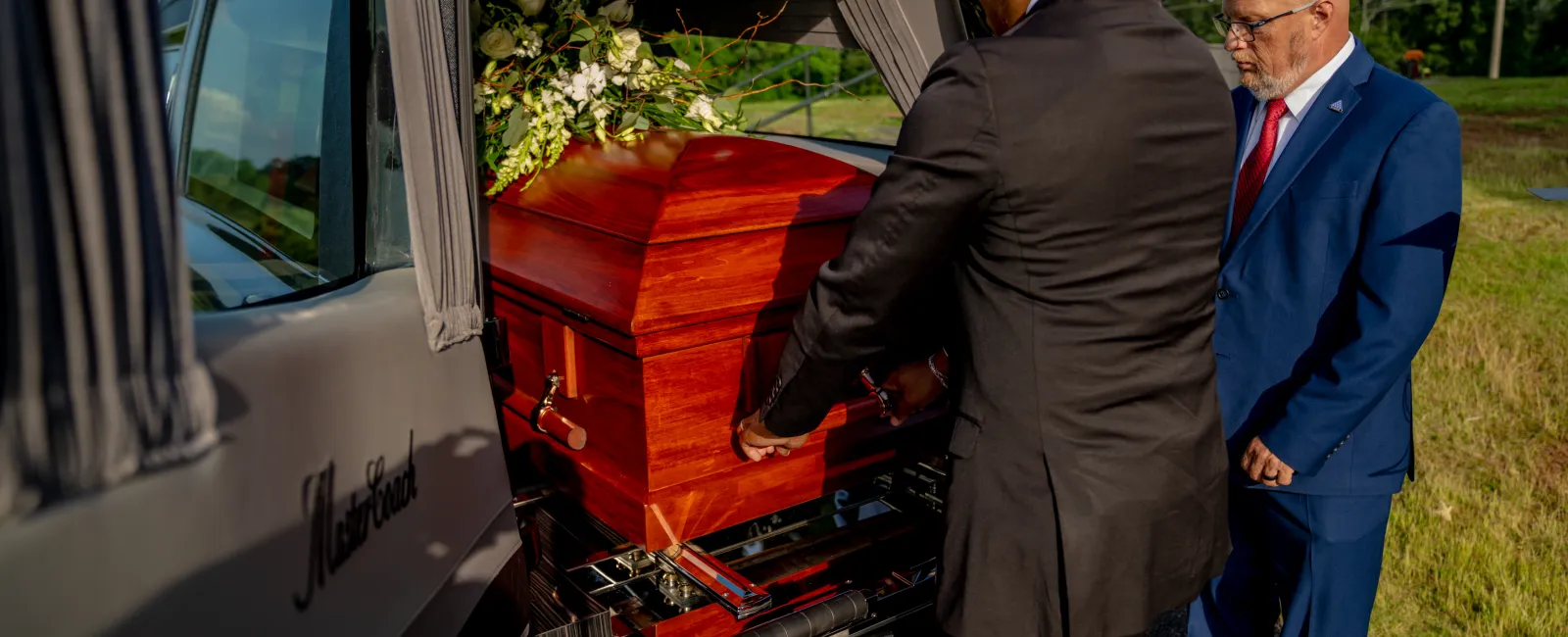 Two men in suits loading a wooden casket with flowers into a funeral vehicle on a sunny day