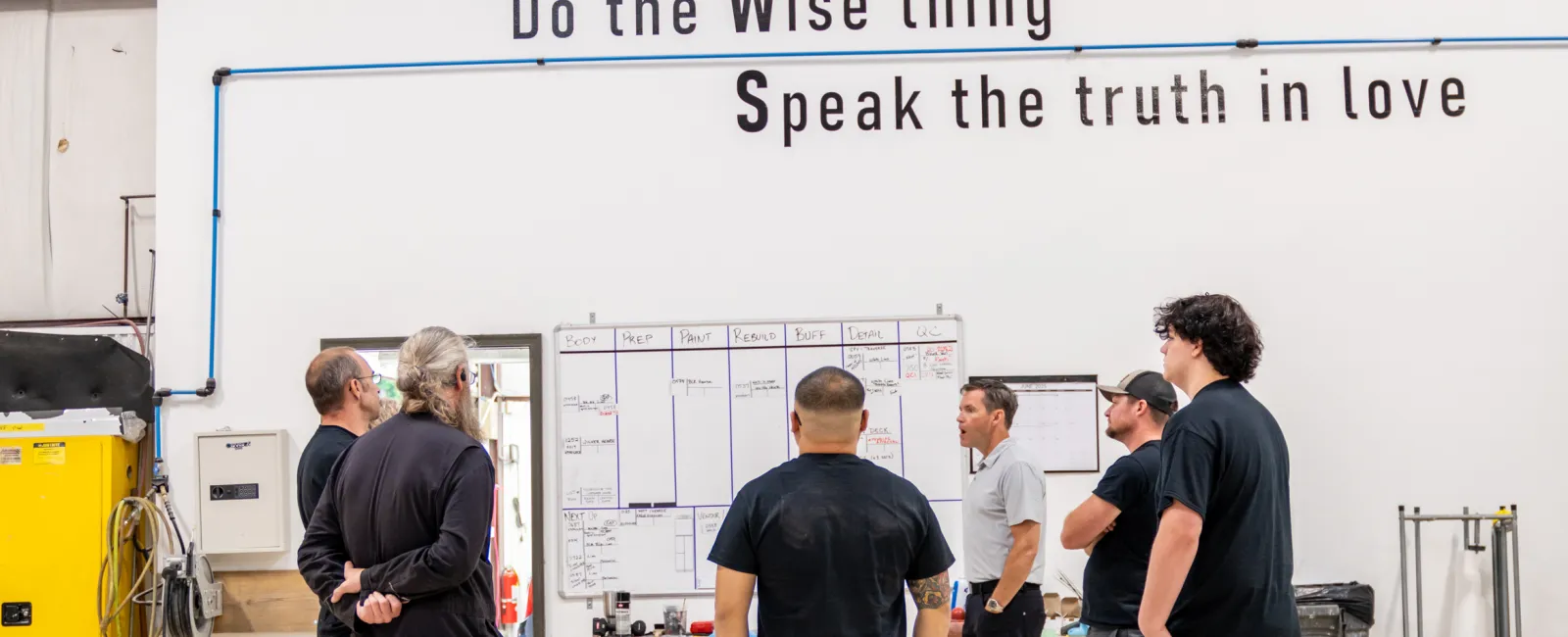 Group of workers standing in a warehouse with motivational words on the wall promoting teamwork and improvement.