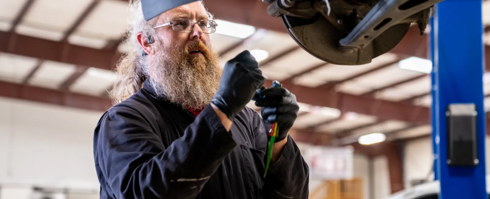 Mechanic with beard and gloves inspecting car underside in a well-lit auto repair shop.