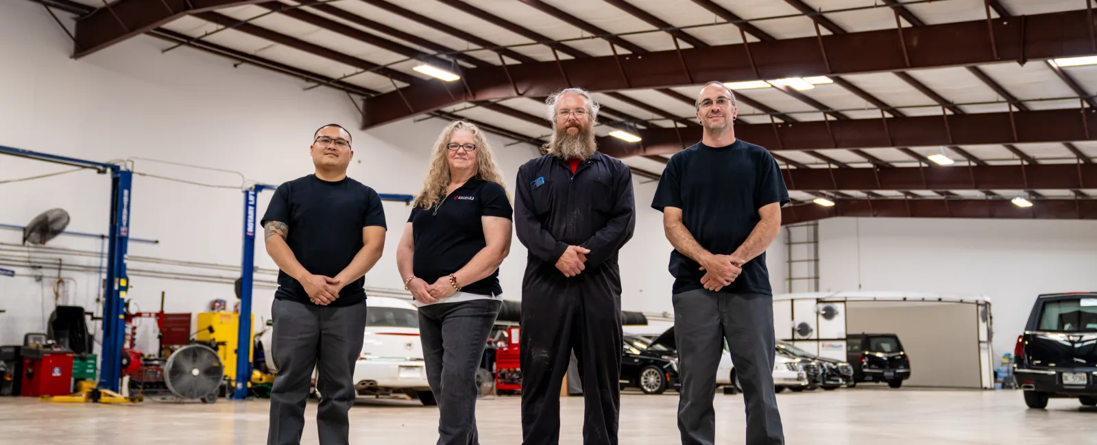 Four automotive technicians standing confidently in a spacious garage with cars and repair equipment in the background.